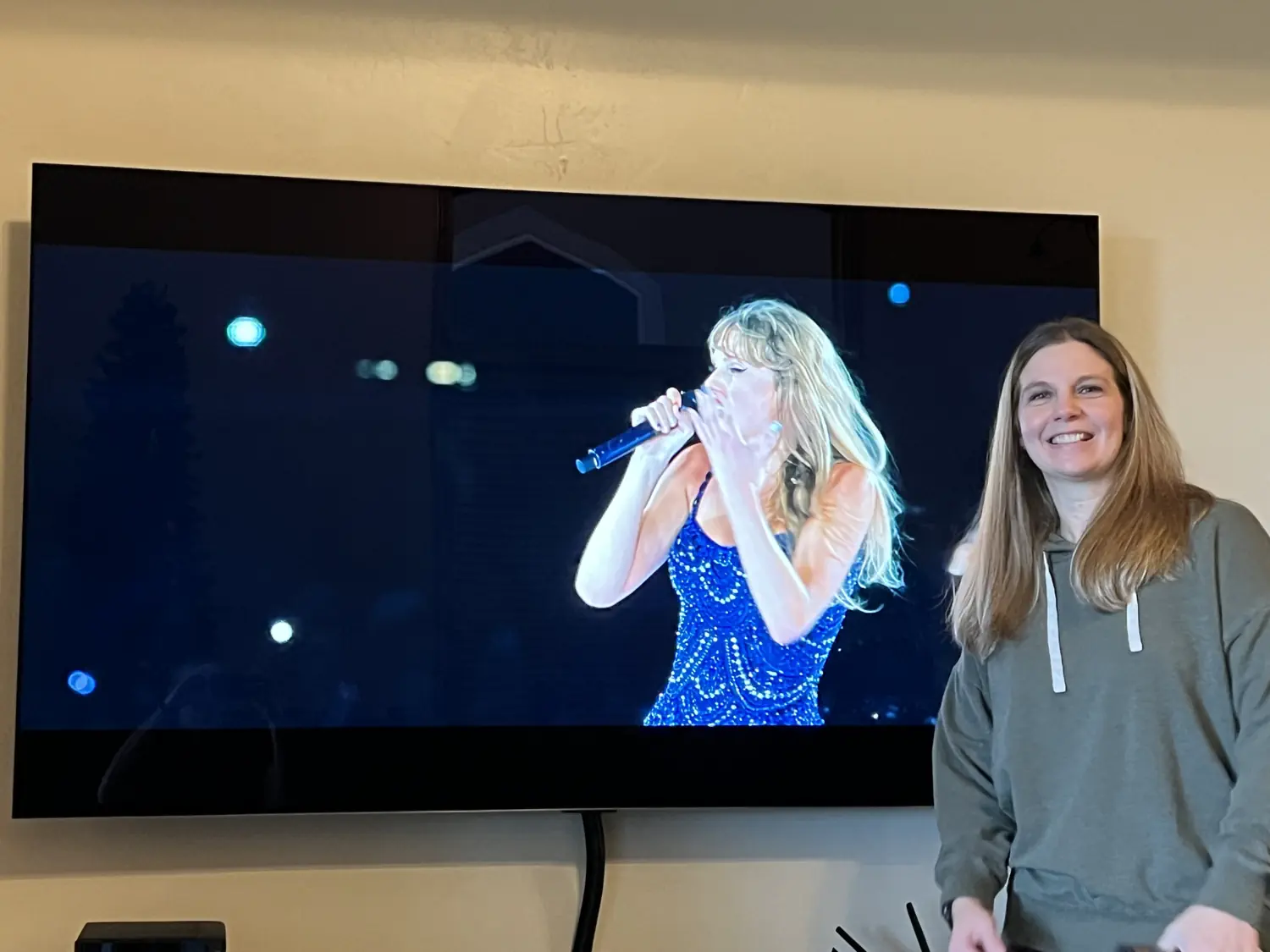 Lindsey is posing in front of a TV displaying Taylor Swift singing on stage